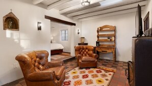 A room with a bed, two leather armchairs, a wooden shelf, a television, and a patterned rug.