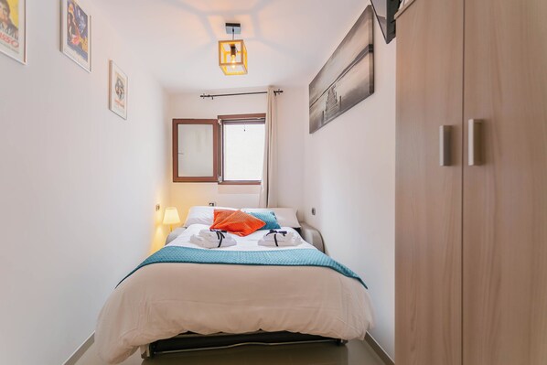 Una cama bien hecha con una colcha verde azulada, un almohadón blanco y uno naranja. A la derecha se ve un armario de madera y, a la izquierda, una ventana con cortinas.