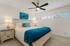 A bedroom with a bed, nightstands, a ceiling fan, a painting of a sea turtle, and windows with blinds.