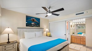 A hotel room with a bed, two bedside lamps, a ceiling fan, a painting of a sea turtle, a bathroom with a vanity and mirror, and a door leading to another room. - Sugar Beach Resort, #409 1 Bedroom Condo by RedAwning (Kihei)