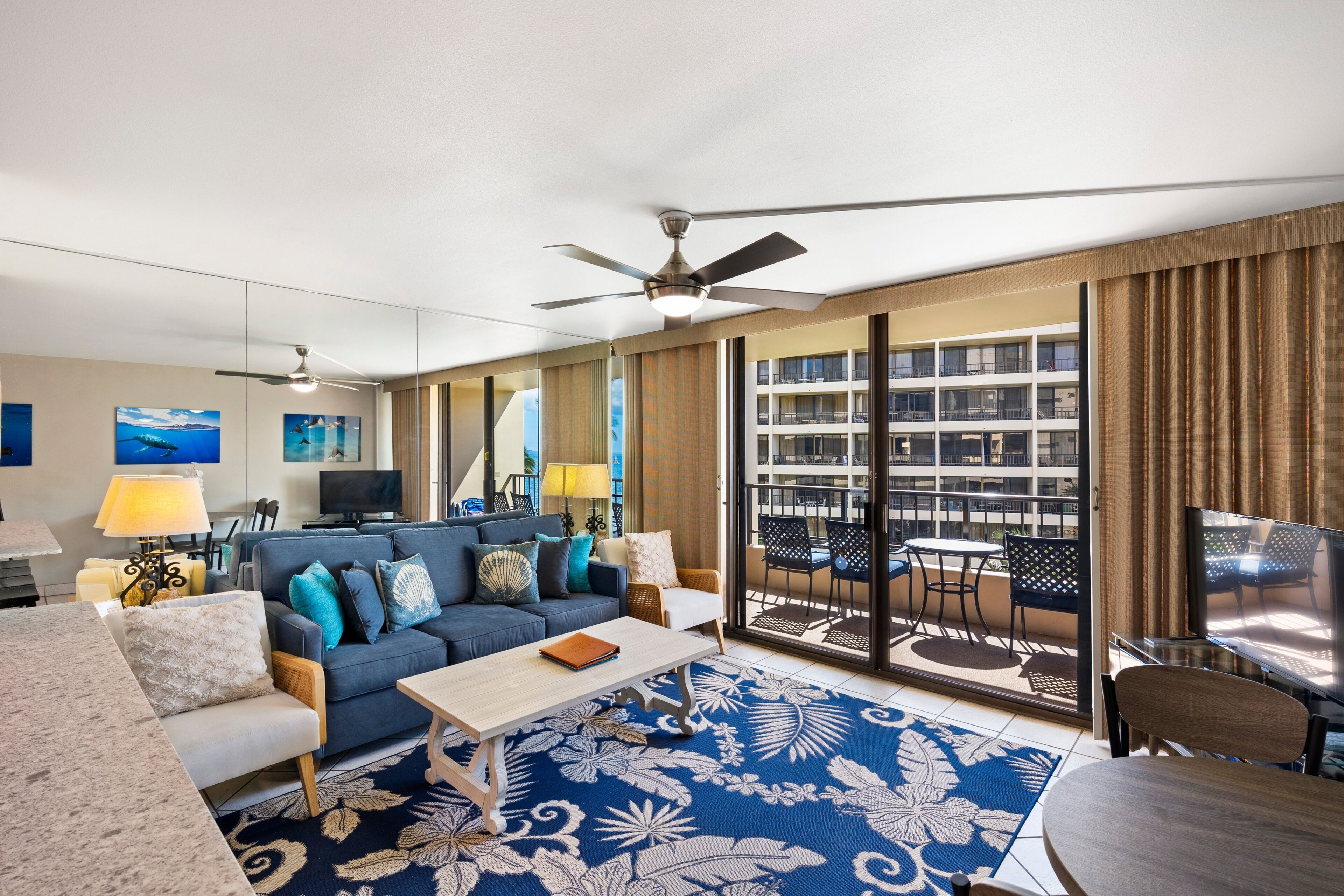A hotel room with a sofa, a coffee table, a TV, a ceiling fan, and a balcony with a table and chairs.