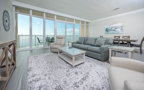 A spacious living room with a large window, a grey sofa, a coffee table, and a dining area with chairs. - Phoenix Gulf Towers 2606 2 Bedroom Condo (Orange Beach)