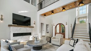 A spacious living area with a flat-screen TV mounted on the wall, a round coffee table, and a staircase leading to an upper level. - Luxury Seagrove w Pool, Hot Tub, Golf Cart & Bikes, Elevator (Seagrove Beach)