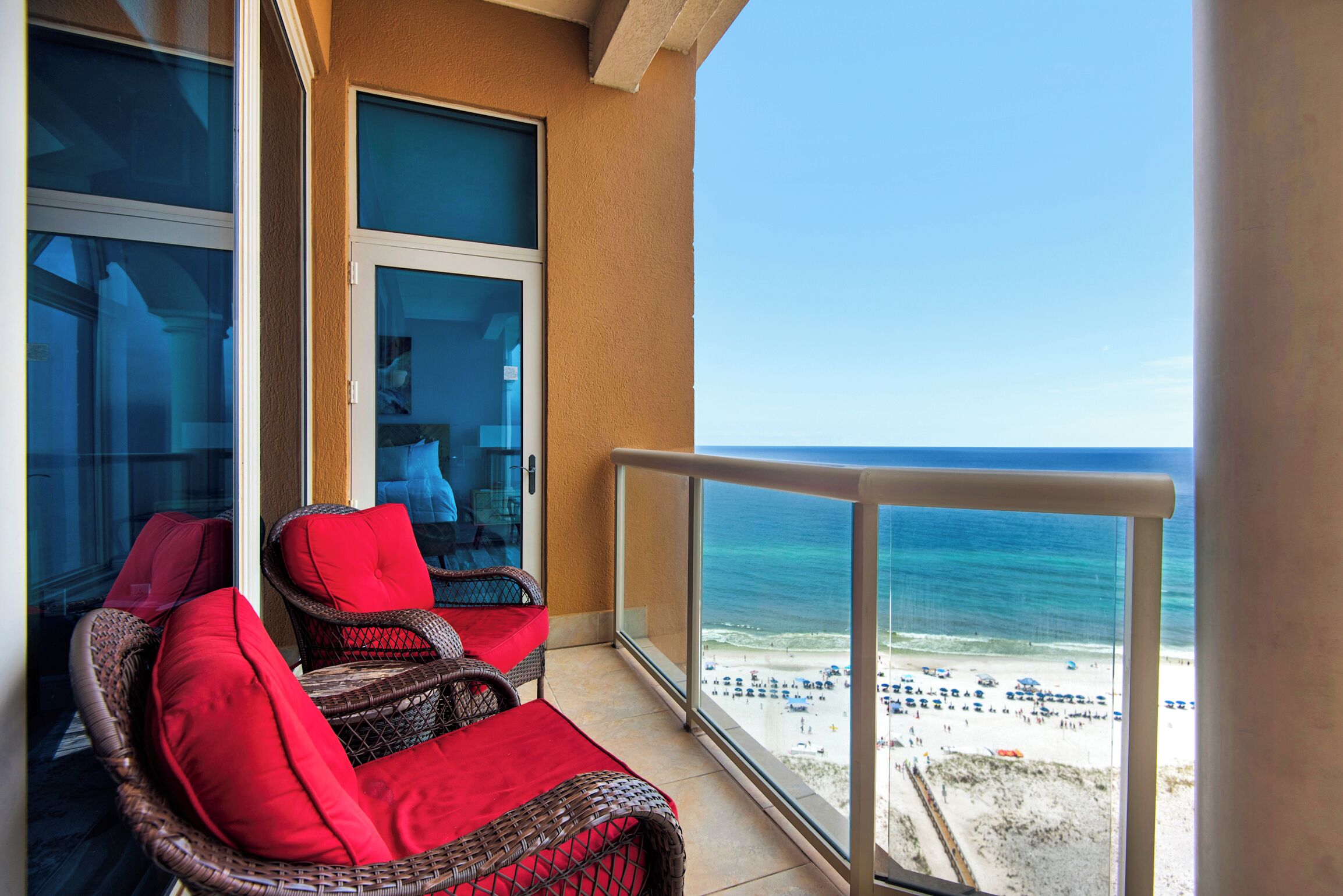 A balcony with wicker chairs and red cushions, overlooking a beach with a clear view of the ocean and beachgoers.