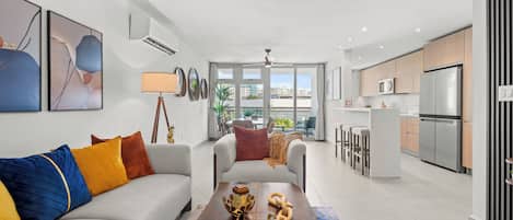Una sala moderna con sofá, una mesa de centro y elementos decorativos. Incluye una zona de cocina con electrodomésticos y una zona de comedor con mesa y sillas.