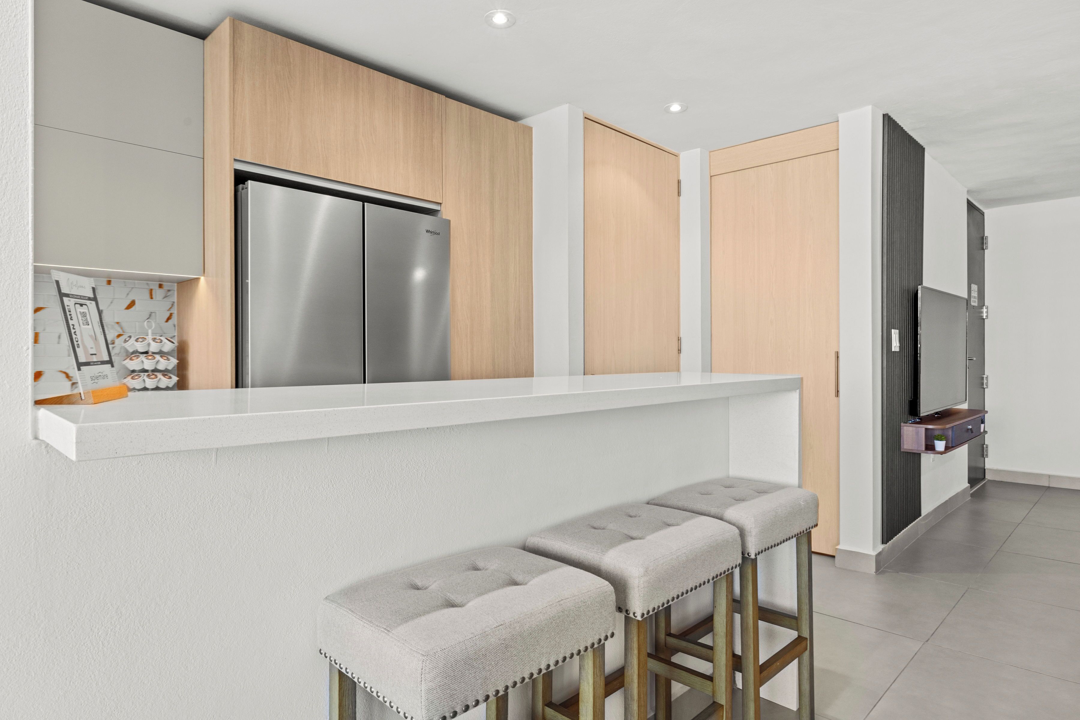 Cocina moderna con refrigerador de acero inoxidable, armarios de madera y encimera blanca. Hay dos taburetes de bar con asientos acolchados.