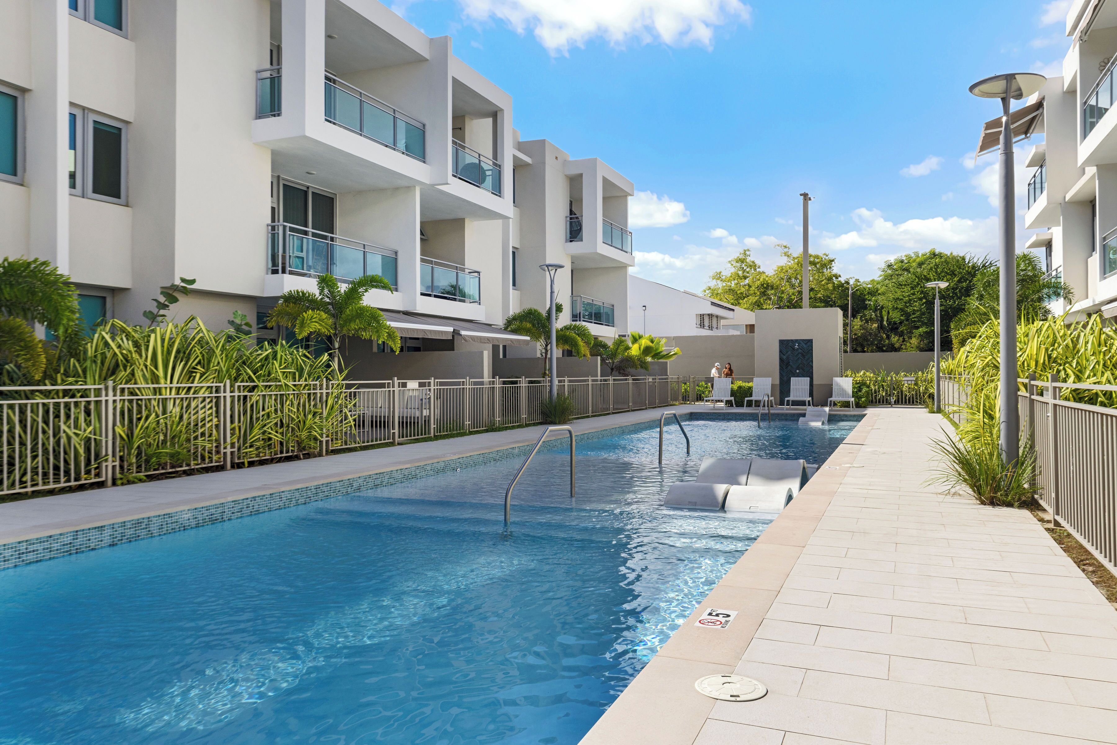 A swimming pool with a tiled deck, surrounded by modern buildings with balconies and a clear sky.