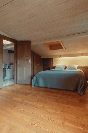 Een slaapkamer met houten vloer, een bed met een blauwe deken en een houten plafond met zichtbare balken.