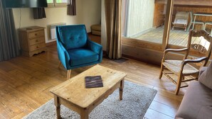 A cozy room with a wooden floor, a blue armchair, a wooden table with a book, a sofa, and a view of a wooden deck through a glass door. - 71m² apartment near Megève, 180m from 🚠 La Princesse, parking, ski room (Demi-Quartier)