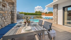 Uma área de refeições ao ar livre com parede de pedra, piscina e cadeiras de descanso ao fundo.