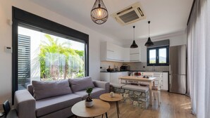 Een modern appartement met een zithoek met een bank, twee ronde tafels en een potplant. De keuken heeft witte kasten, een eettafel en een koelkast.