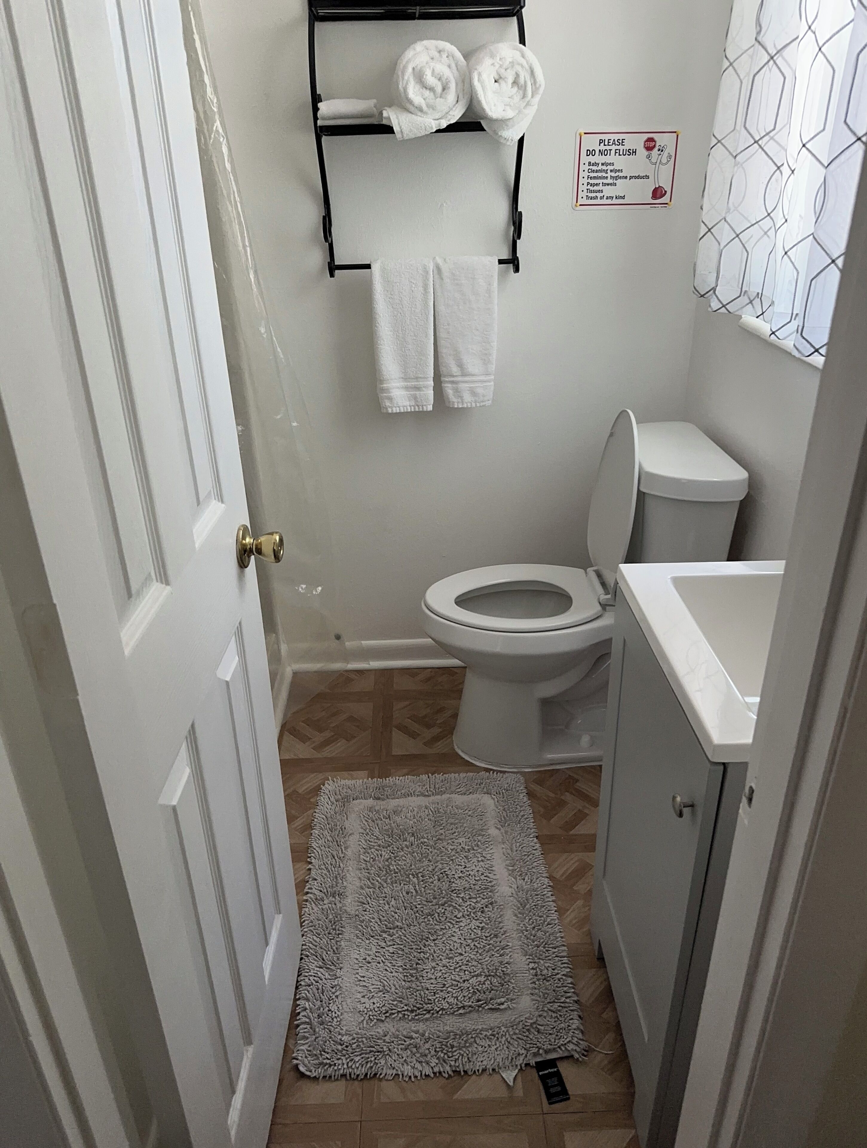 A small bathroom with a toilet, a towel rack with two towels, a sign prohibiting flushing of certain items, and a patterned shower curtain.