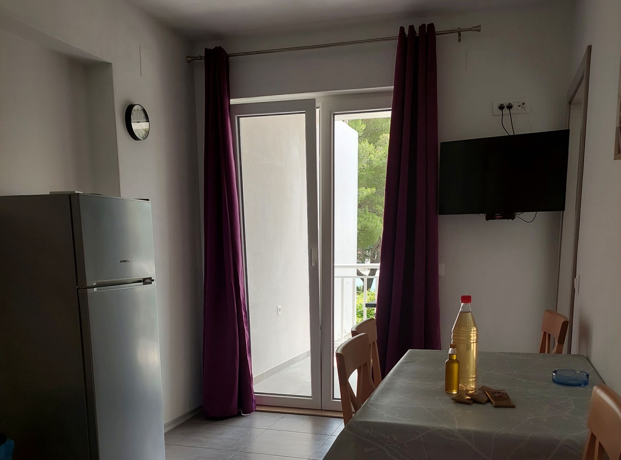 Une petite cuisine équipée d’un réfrigérateur, d’une table à manger avec une bouteille et un cendrier, et d’un téléviseur fixé au mur.