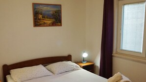 A bedroom with a bed, a nightstand with a lamp, a painting on the wall, and a window with blinds. - Beach House Žuljana (Ston)
