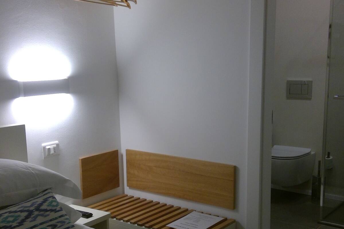 Habitación de hotel con cama, cabecero de madera, mesita de noche con lámpara, perchero y vista al baño.