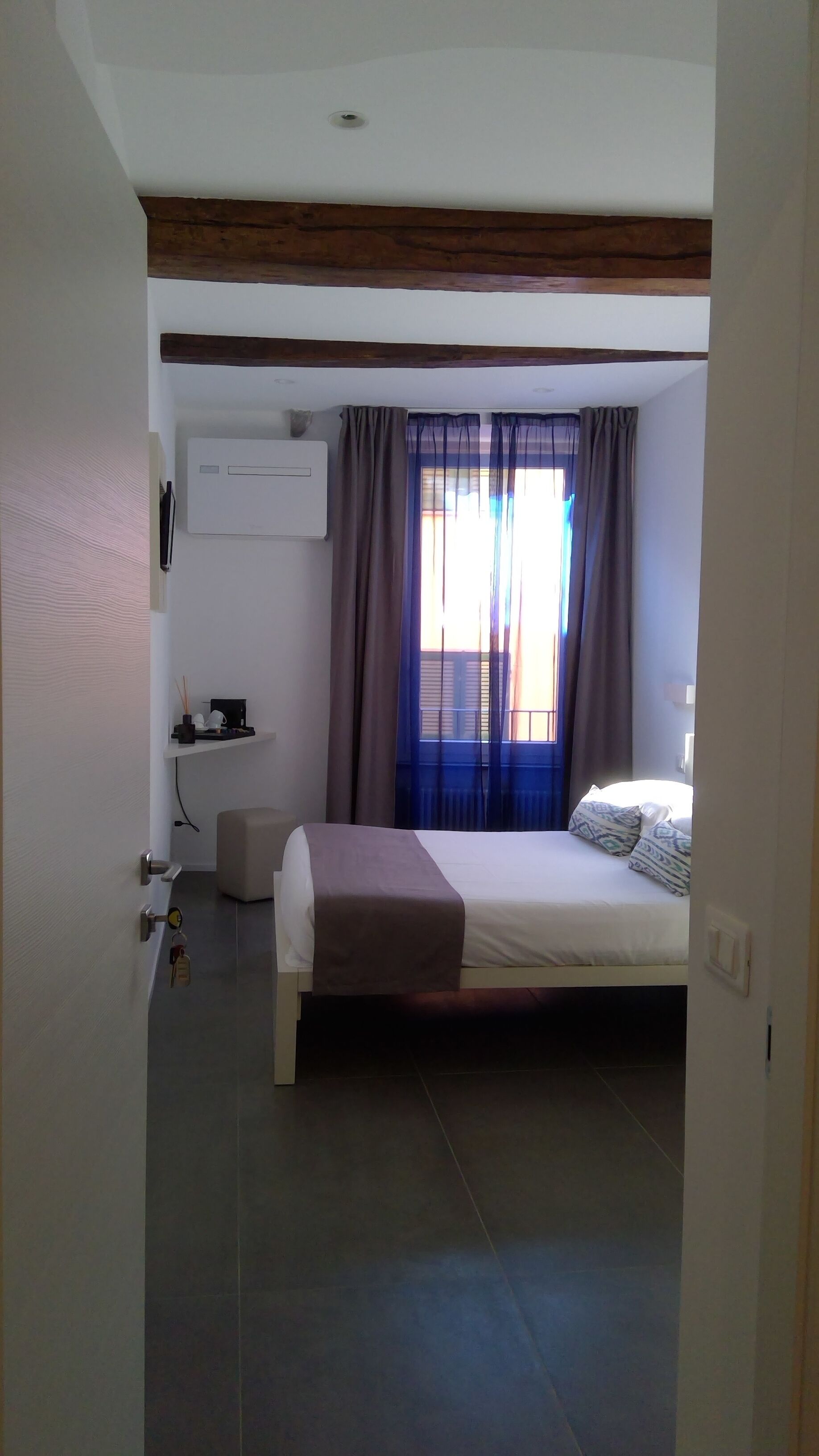 A bedroom with a bed, a bedside table, a wall-mounted air conditioner, and a window with curtains.