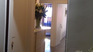 A hallway with a door on the left, a vase with flowers on a counter, and a view into another room with a window. - OBLO' PORTOVENERE GUESTHOUSE (Portovenere)