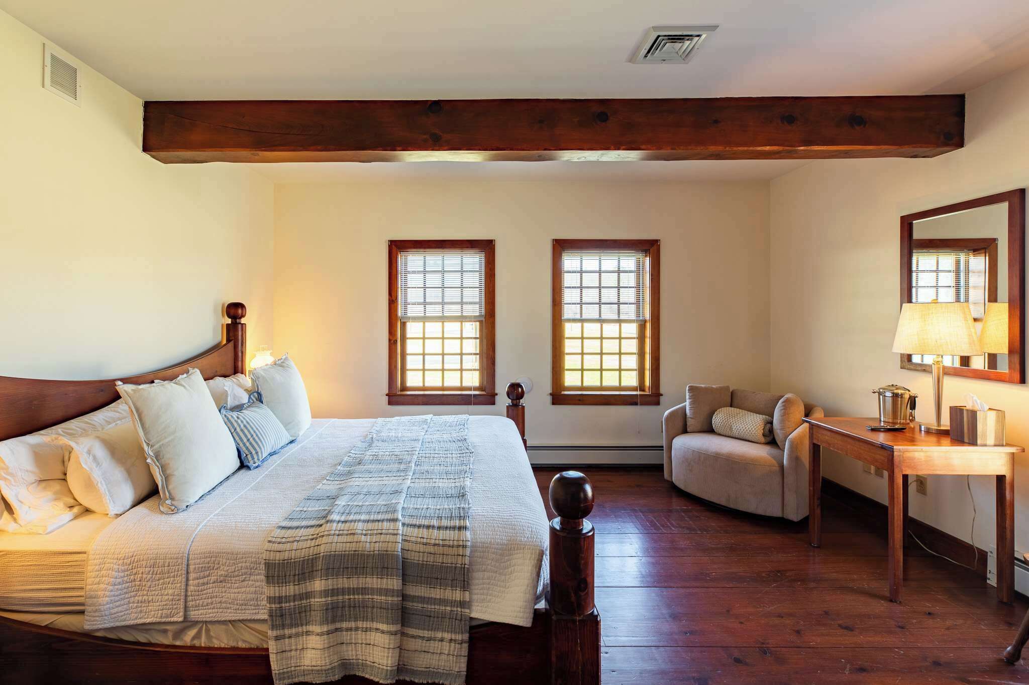A bedroom with a large bed, two windows, a chair, a table, and a mirror.