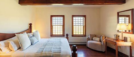 A bedroom with a large bed, two windows, a chair, a table, and a mirror.