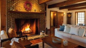 A cozy living room with a brick fireplace, wooden beams, a white sofa, and a clock on the wall.