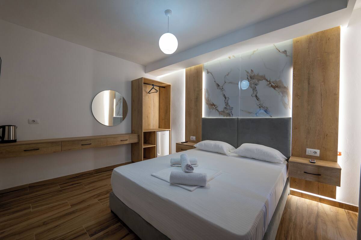 Una habitación de hotel moderna con una cama grande, suelo de madera, un armario de madera, una mesita de noche y un espejo fijado a la pared.