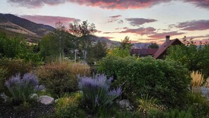 A house with a red roof nestled in a lush garden with various plants and a mountainous backdrop.