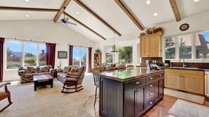 A spacious living area with a large island kitchen, wooden beams, and large windows offering views of the outdoors.