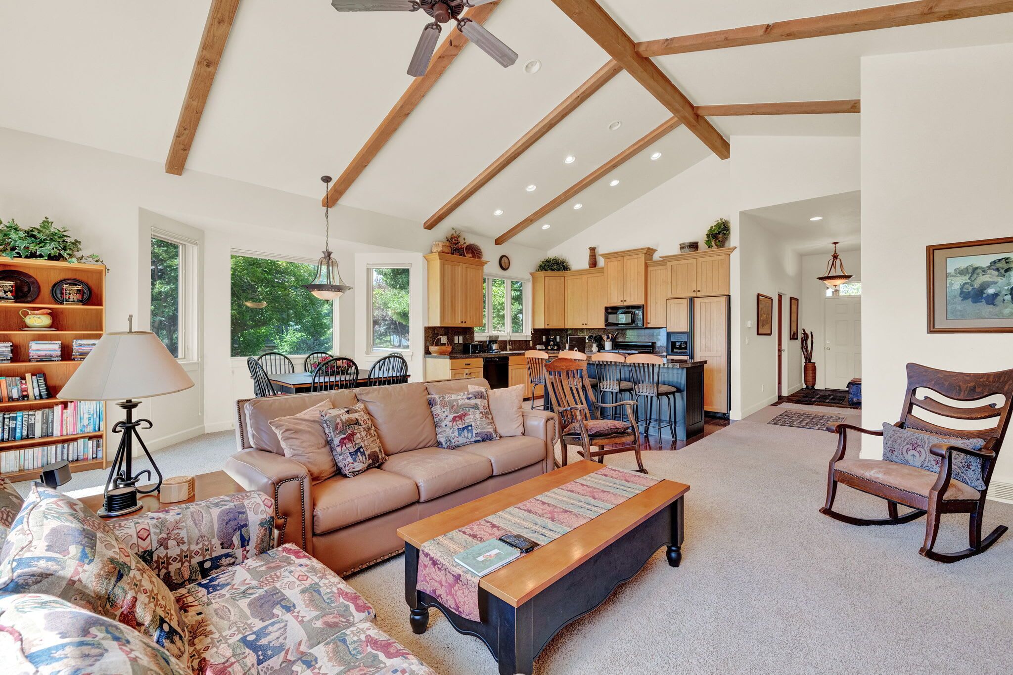 A spacious living room with a sofa, coffee table, and rocking chair. A kitchen is visible in the background with wooden cabinets and a dining area.