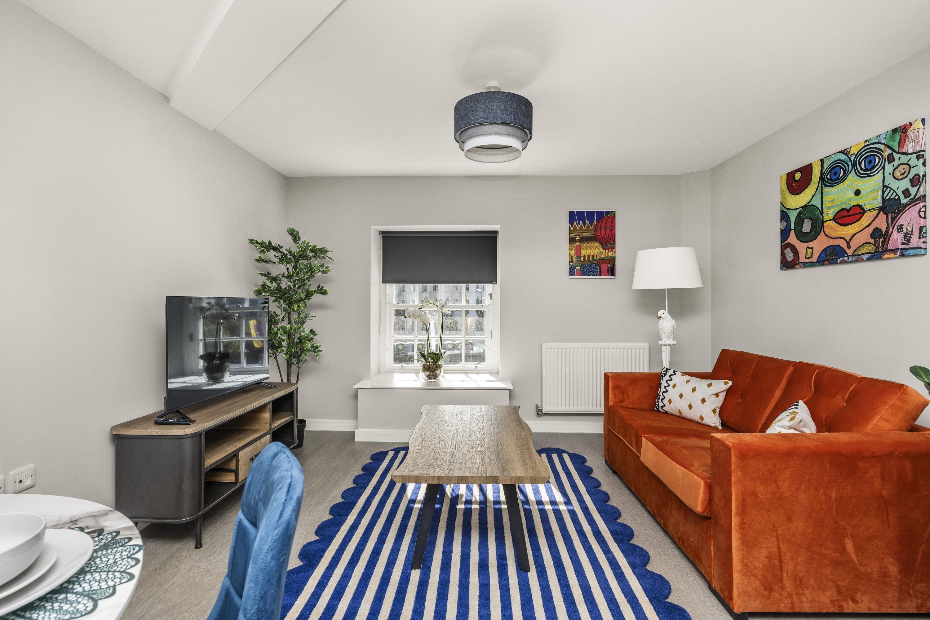 Una sala de estar moderna con una mesa de centro de madera, un tapete azul y blanco a rayas, un sofá naranja, un televisor sobre un mueble de madera y una ventana con vista a la ciudad.