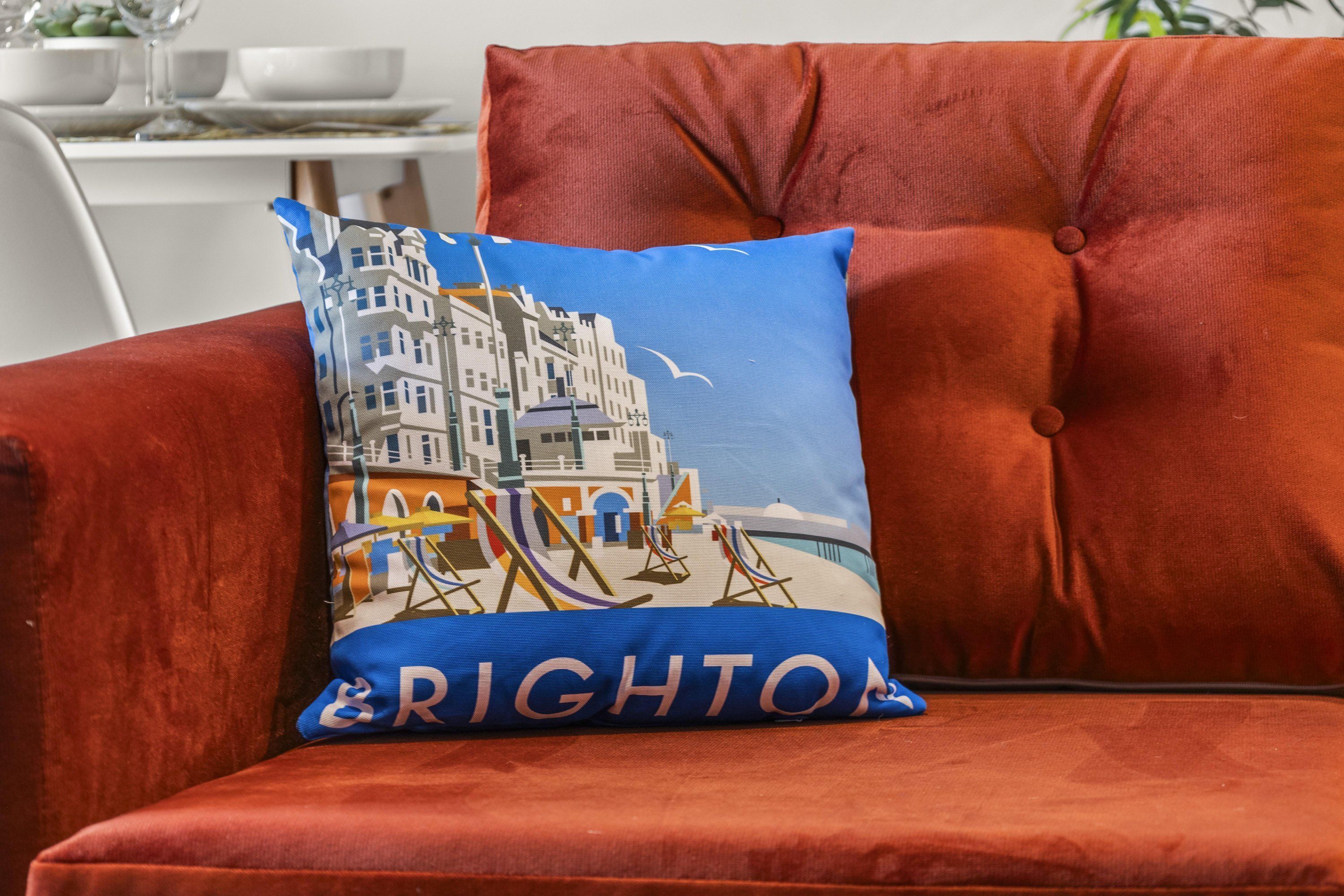 A red velvet sofa with a blue pillow featuring a seaside town illustration and the word "BRIGHTON".