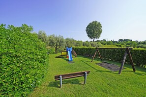 Un'area verde con prato, un'area giochi, una panchina e un albero.