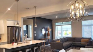 A modern kitchen with a central island, bar stools, a dining table, a gray sofa, and a large window.