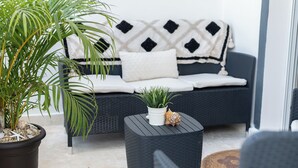 Una sala moderna con un sofá azul oscuro, cojines blancos y una manta estampada. Hay una planta grande en maceta y una mesita con un objeto decorativo.