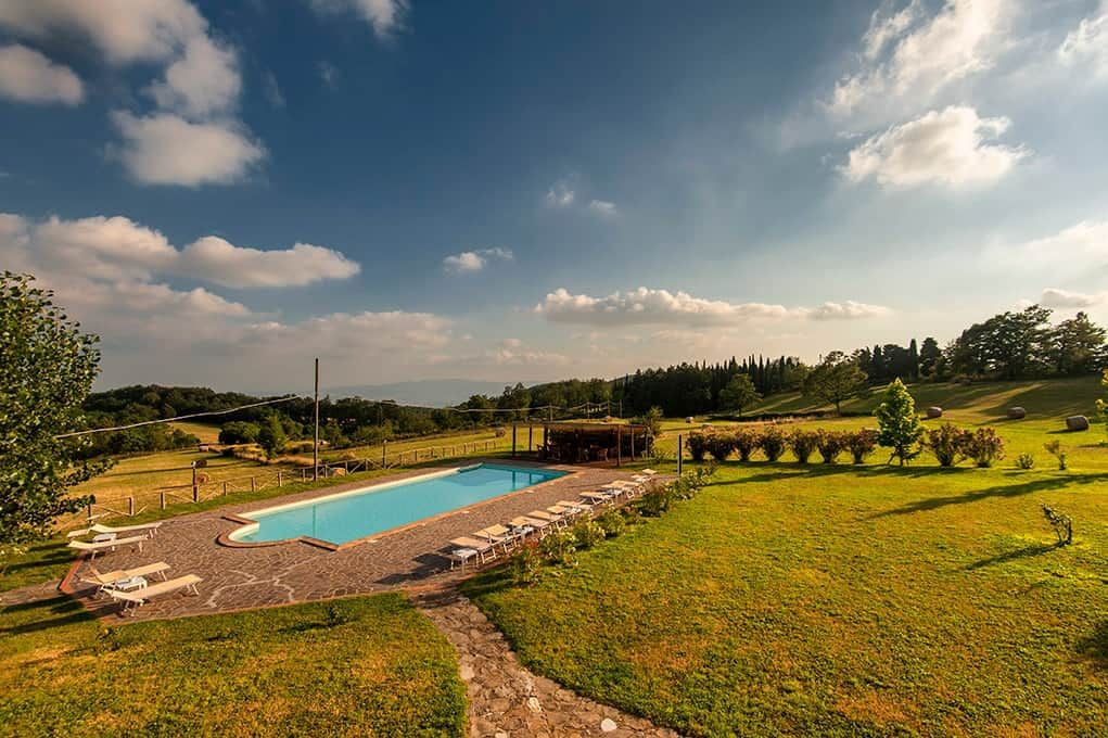 A swimming pool with lounge chairs and a covered area, surrounded by a grassy landscape and trees.
