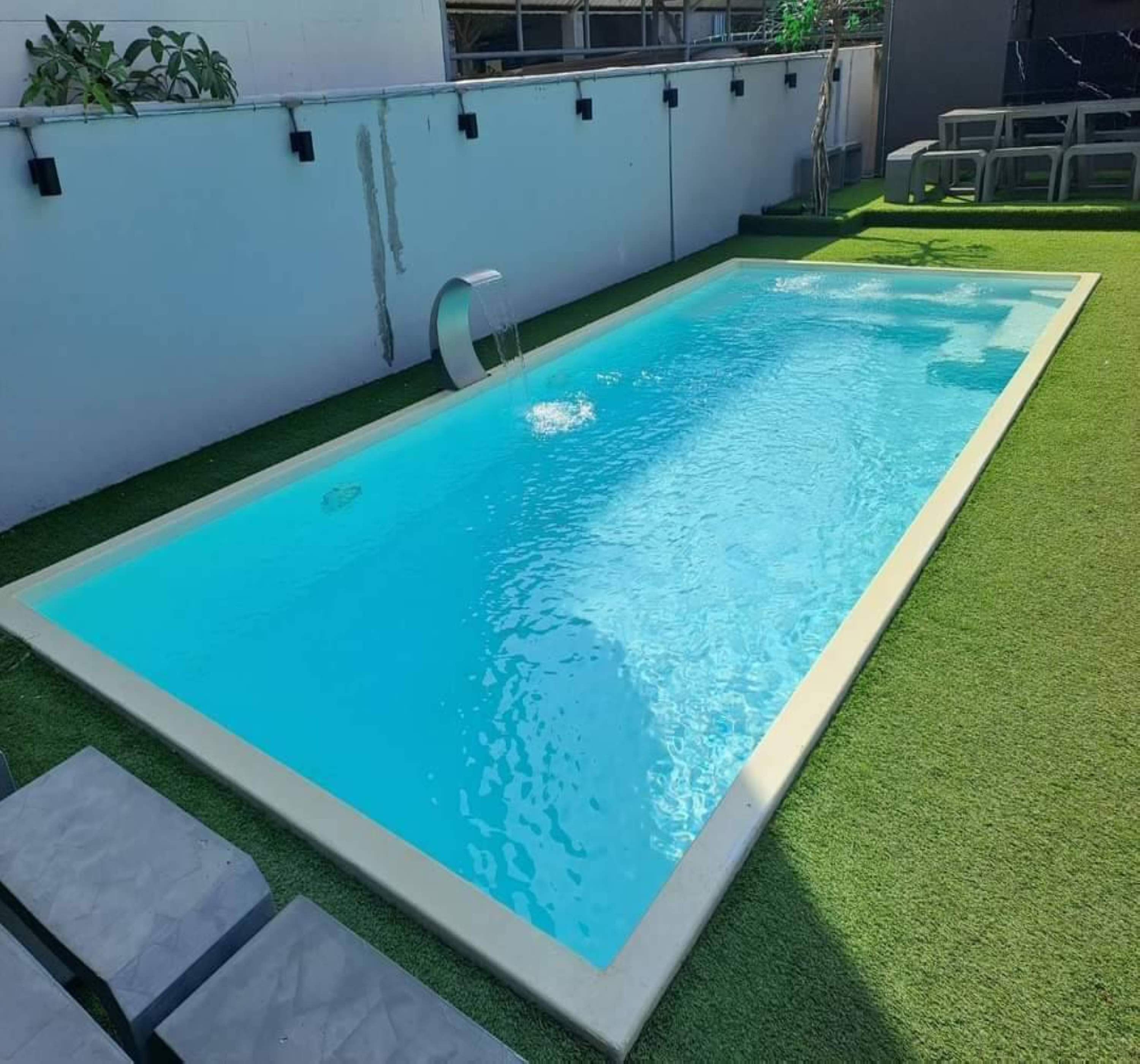 A rectangular swimming pool with a clear blue water surface, surrounded by artificial green grass and a white wall with black fixtures.