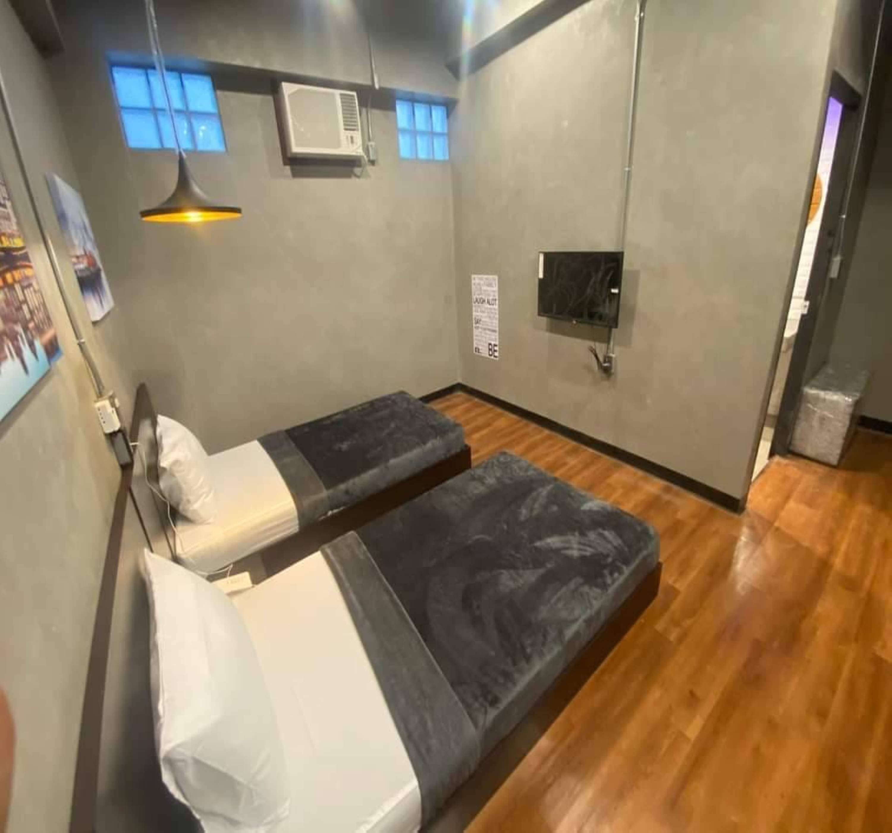 Una habitación pequeña y funcional con dos camas individuales, aire acondicionado de pared, un televisor pequeño y una ventana con persianas.