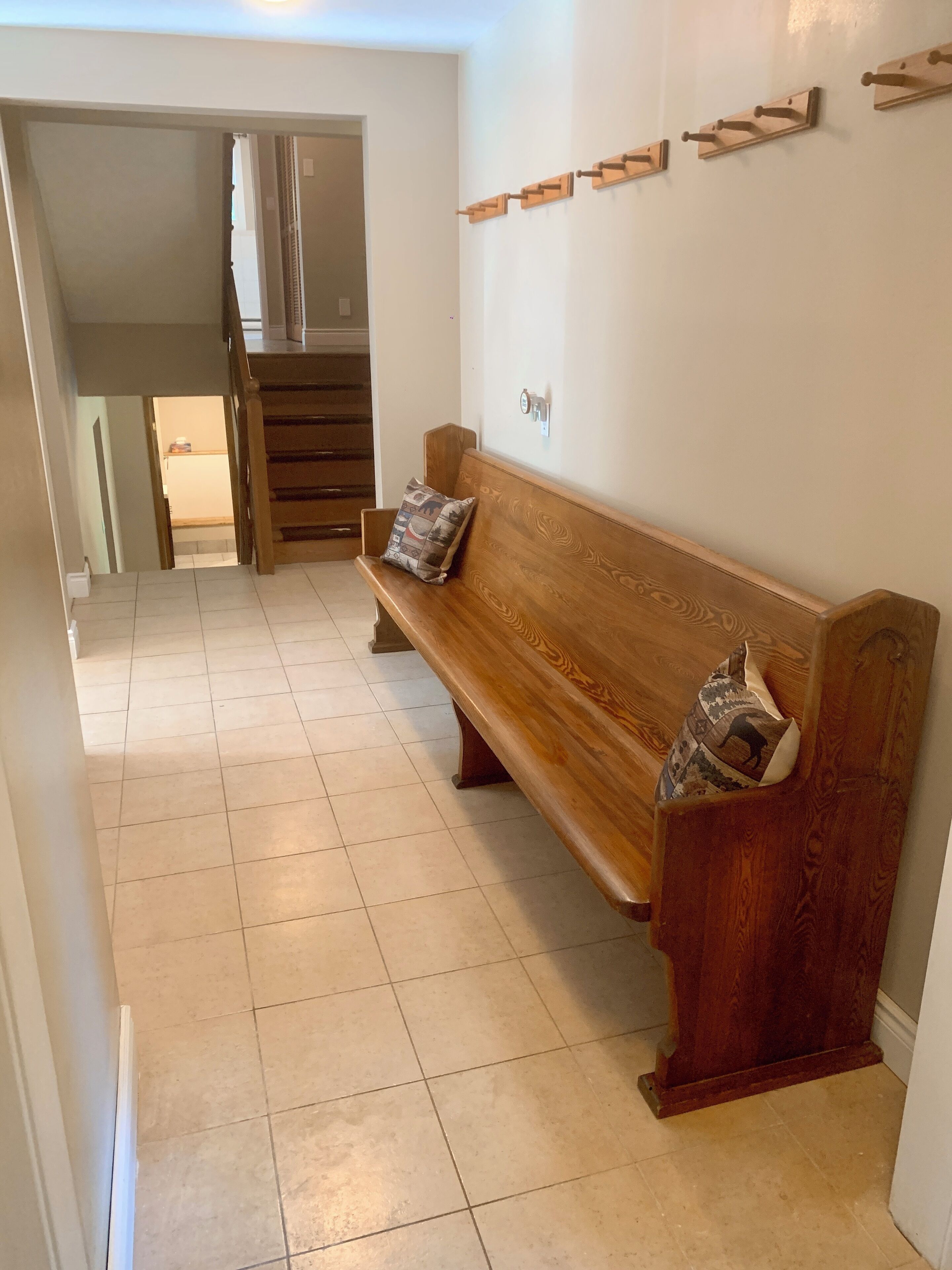 Un banco de madera con cojines estampados en un pasillo con suelo de baldosas, una escalera de madera e interruptor de luz instalado en la pared.