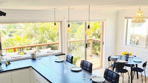 A modern kitchen with a large island, dining area with chairs, and a view of greenery through large windows. - Treeview - A Beautiful 3-bed House in Kiama (Kiama)
