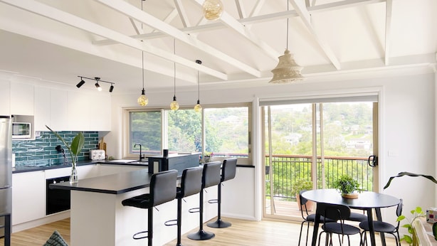 A modern kitchen with a central island, black bar stools, and a dining area with a round table and chairs. - Treeview - A Beautiful 3-bed House in Kiama (Kiama)