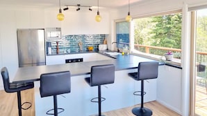 A modern kitchen with a central island, bar stools, and a large window offering a view of the outdoors. - Treeview - A Beautiful 3-bed House in Kiama (Kiama)