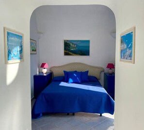 Une chambre à coucher avec un lit, deux tables de chevet, un tableau au mur et une porte donnant sur une autre pièce.