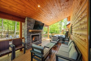 A covered outdoor deck with a stone fireplace, wooden furniture, and a view of the forest.