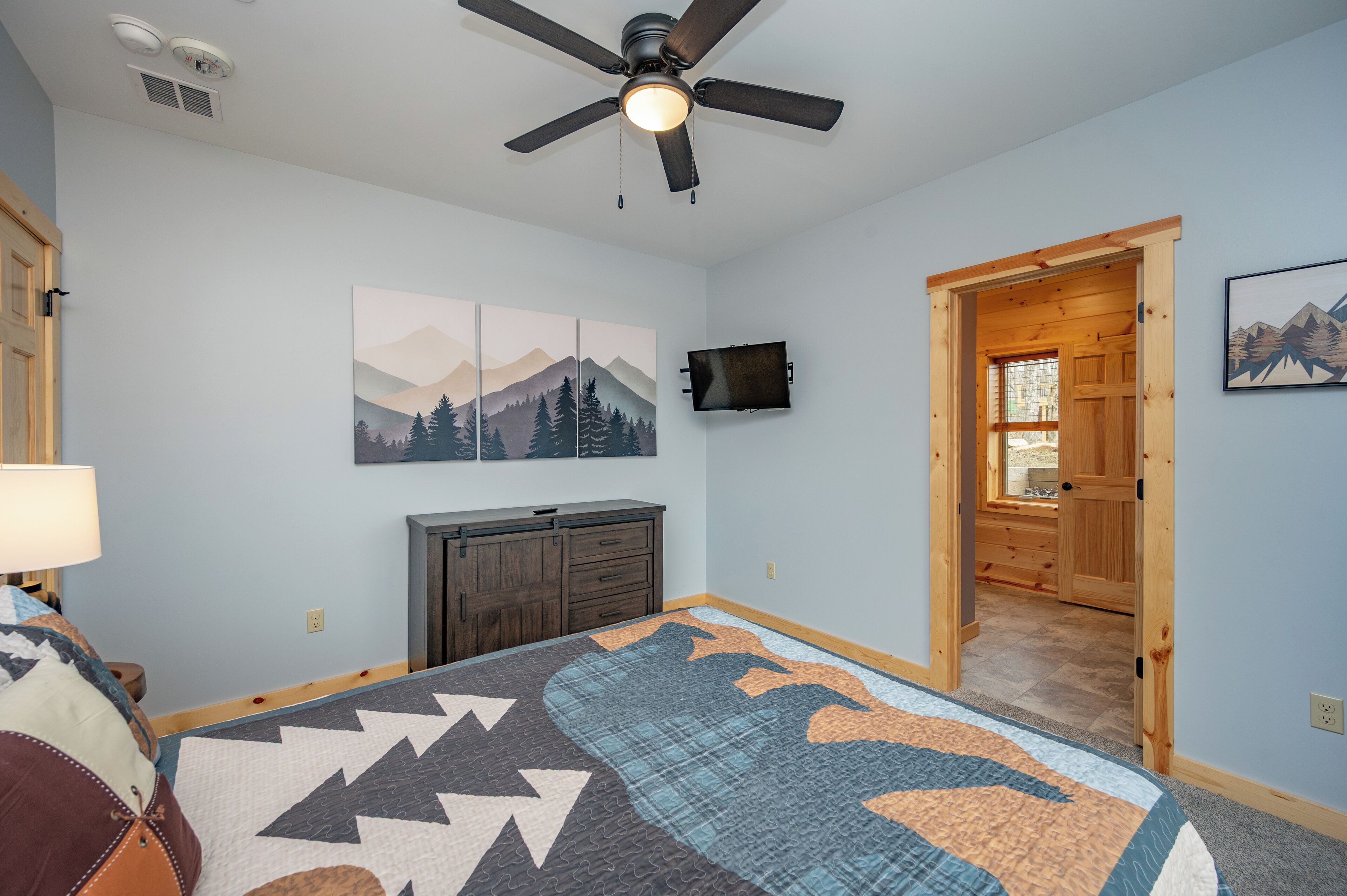 A bedroom with a bed, a ceiling fan, a mounted TV, a wooden door, and a mountain landscape painting.