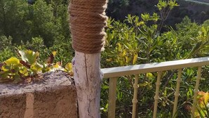 Un balcon doté d’un poteau rustique enroulé de corde, offrant une vue panoramique sur un paysage pittoresque avec des collines et un village en contrebas.