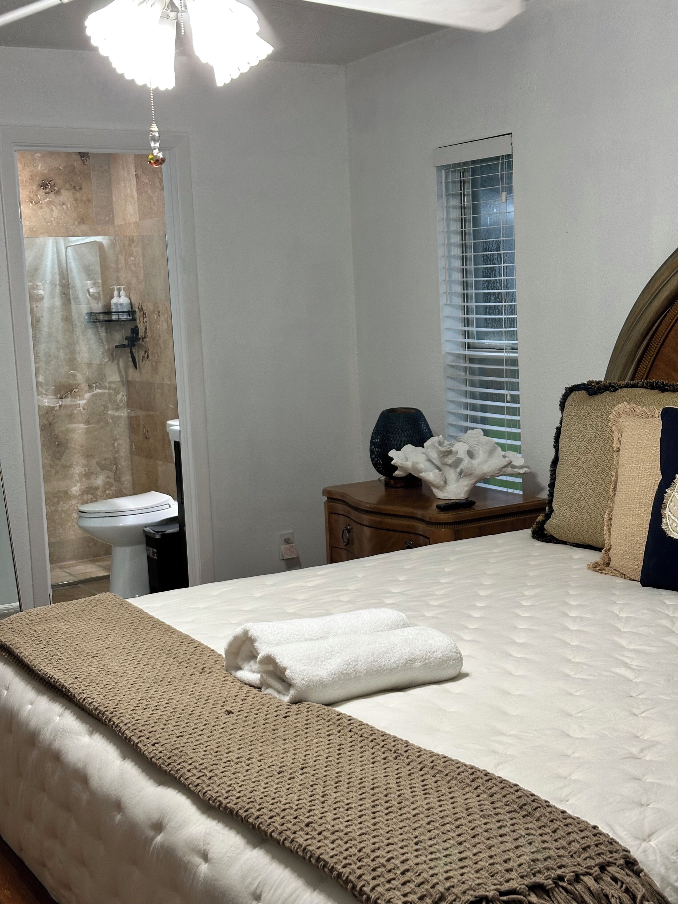A bedroom with a bed, a nightstand, a window with blinds, a shower area, and a ceiling light.