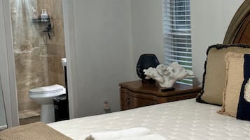 A bedroom with a bed, a nightstand, a window with blinds, a shower area, and a ceiling light.