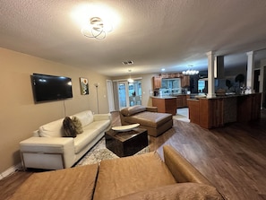 A living room with a white sofa, a brown leather couch, and a coffee table. There is a kitchen area with wooden cabinets and a countertop.