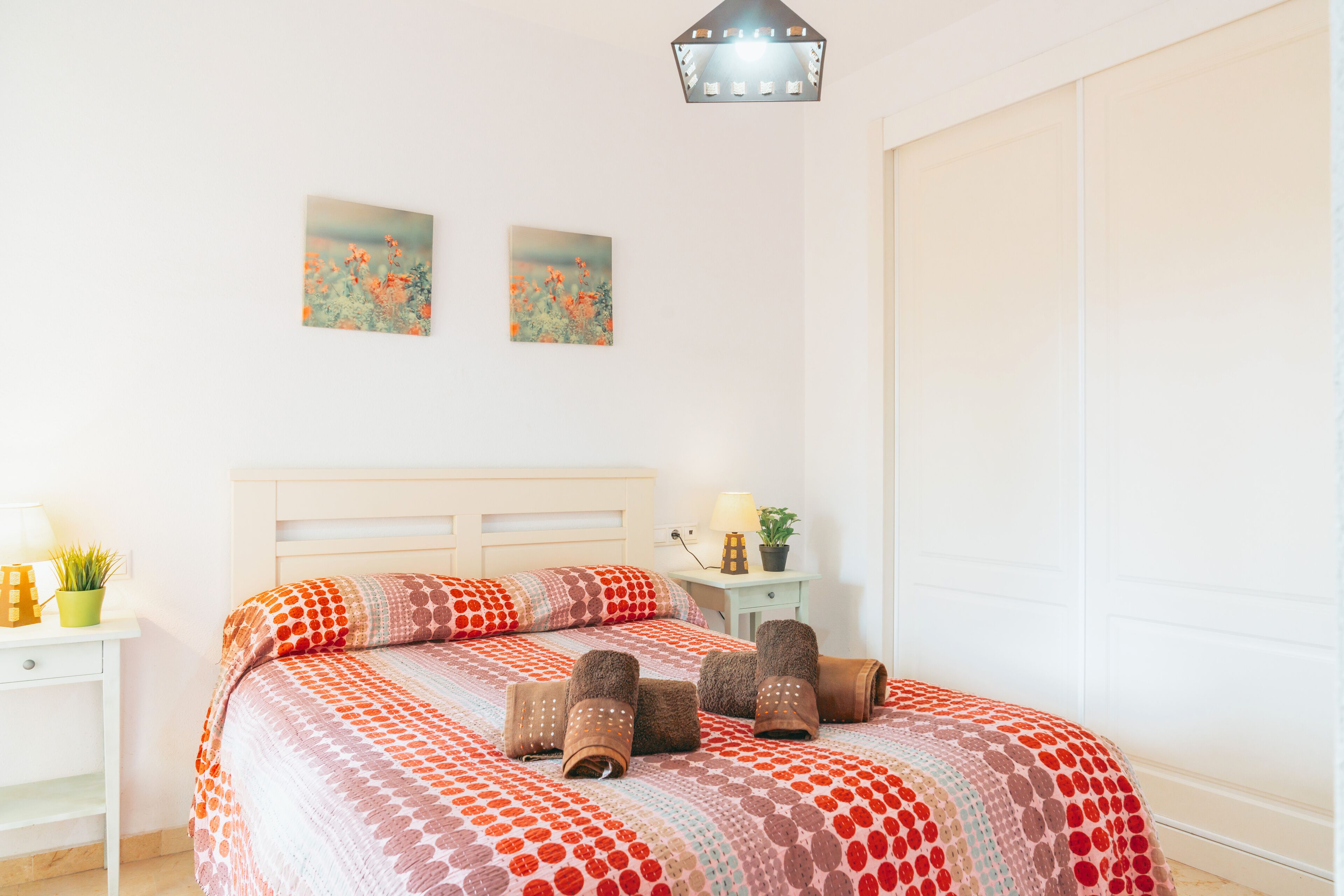 A neatly made bed with a patterned blanket, a bedside table with a lamp and a plant, and two wall-mounted pictures.