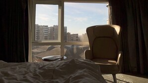 A hotel room with a bed, a chair, and a large window offering a view of buildings. - Luxe Apartment - Stylish 2 Bedroom Apartment (Harrow)