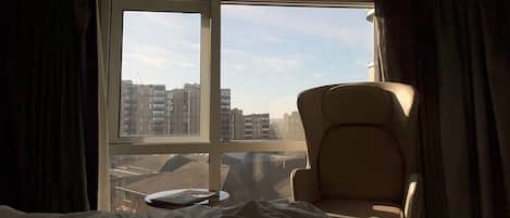 Hotellihuone, jossa on sänky, tuoli ja suuri ikkuna, josta avautuu näkymä rakennuksiin.